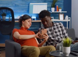 Married interracial couple relaxing on living room sofa at home. Pregnant caucasian woman using smartphone while african american man watching movie on television with remote control
