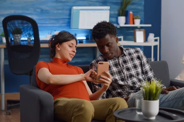Married interracial couple relaxing on living room sofa Married interracial couple relaxing on living room sofa at home. Pregnant caucasian woman using smartphone while african american man watching movie on television with remote control