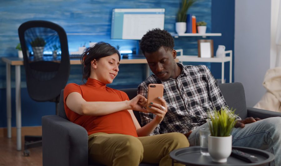 Married interracial couple relaxing on living room sofa at home. Pregnant caucasian woman using smartphone while african american man watching movie on television with remote control