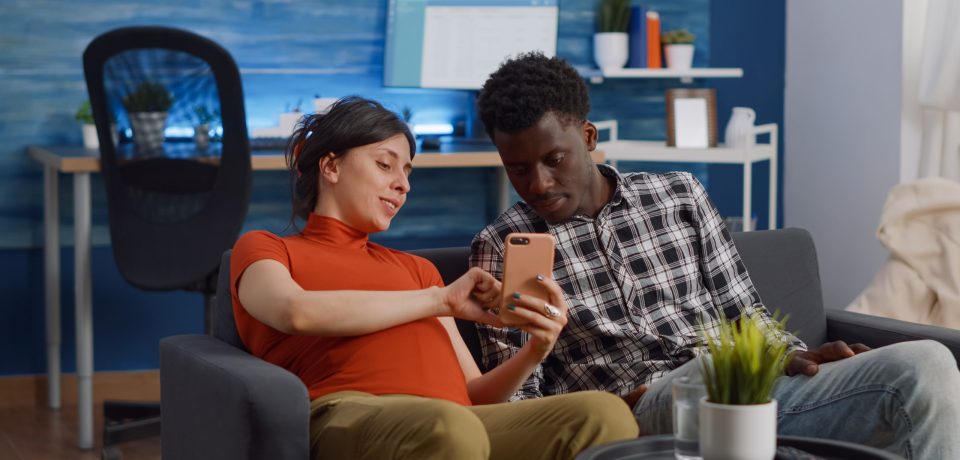 Married interracial couple relaxing on living room sofa at home. Pregnant caucasian woman using smartphone while african american man watching movie on television with remote control